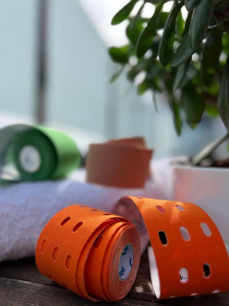 Fotografía con profundidad de campo de rollos de vendaje neuromuscular en primer plano, destacando una cinta naranja perforada. Al fondo, se aprecian rollos verde y beige junto a una planta de interior sobre una mesa de madera.