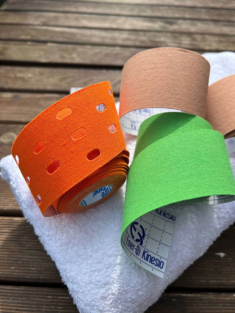 Primer plano cenital de tres rollos de cinta kinesiológica de colores naranja, verde y beige, reposando sobre una toalla blanca en una superficie de madera al aire libre.