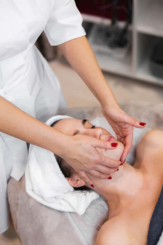 Vista desde arriba de unas manos con uñas rojas tocando el mentón y los pómulos de una persona con el cabello envuelto en una toalla blanca.