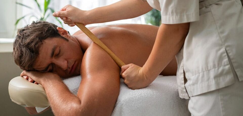 masaje-tailandes-espalda-coruna-natalia-huba Vista lateral de un hombre relajado en una camilla de masajes mientras una profesional realiza una técnica de maderoterapia utilizando una caña de bambú sobre los músculos de la espalda y el hombro en un estudio iluminado.
